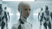 Face of blond haired girl whos is actually an android in a futuristic room with more androids in the background